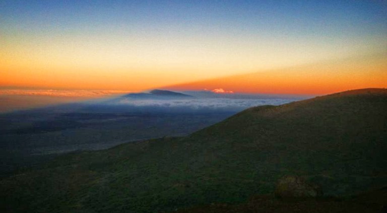 Turning back during sunset. Mt. Hualalai is seen in the distance as a dark blue peak extending above the cloud layer. The diagonal line seen starting in the dark blue shadow on the lefthand side and extending into the red-orange sky is the shadow of Mauna Kea.