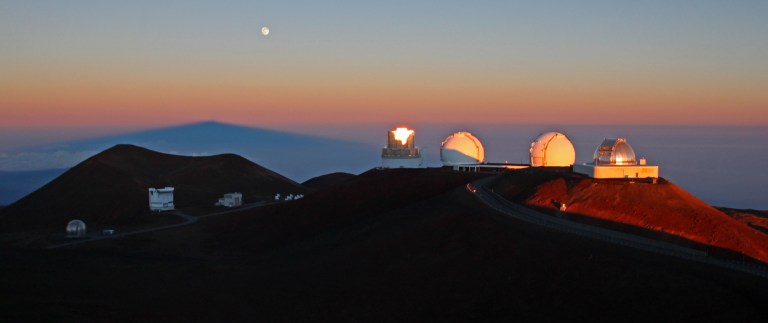 Sunrise over Mauna Kea. Four large telescopes from left to right: Subaru, Keck I and II, IRTF. Lower left are the telescopes that make up submillimeter valley. The dark blue peak in the background is the shadow of Mauna Kea. On full moon nights, the moon sets just as the sun rises.